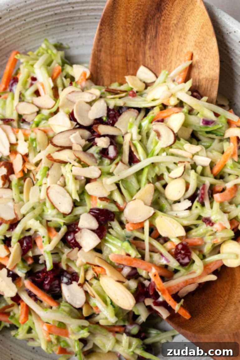 A hand scooping up a generous serving of creamy broccoli slaw from a large bowl, showcasing its inviting texture and readiness to be enjoyed.