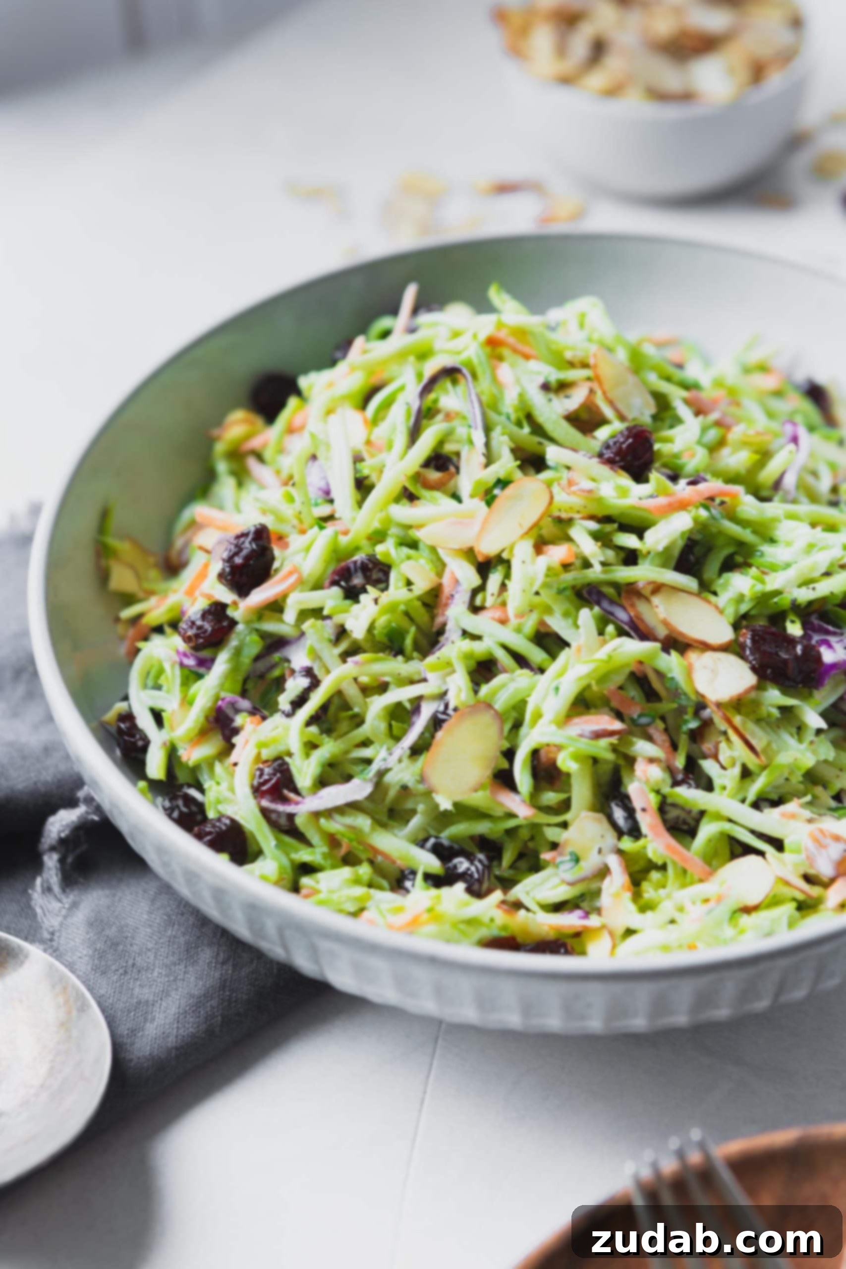 A photo of a big white ceramic bowl full of broccoli slaw topped with raisins and sliced almonds.