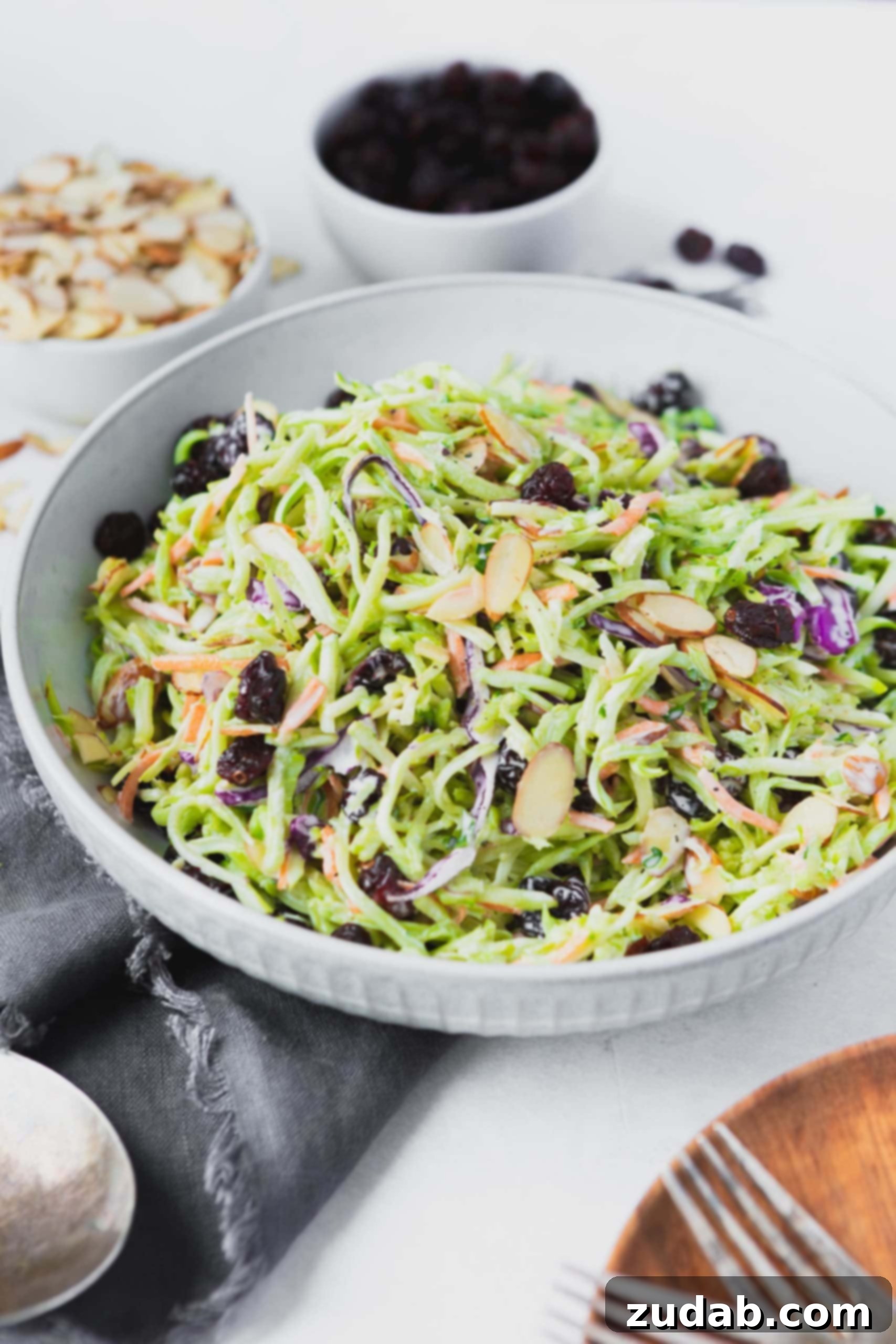 A photo of a bowl full of broccoli slaw topped with sliced almonds and plump raisins, all in a large white bowl.