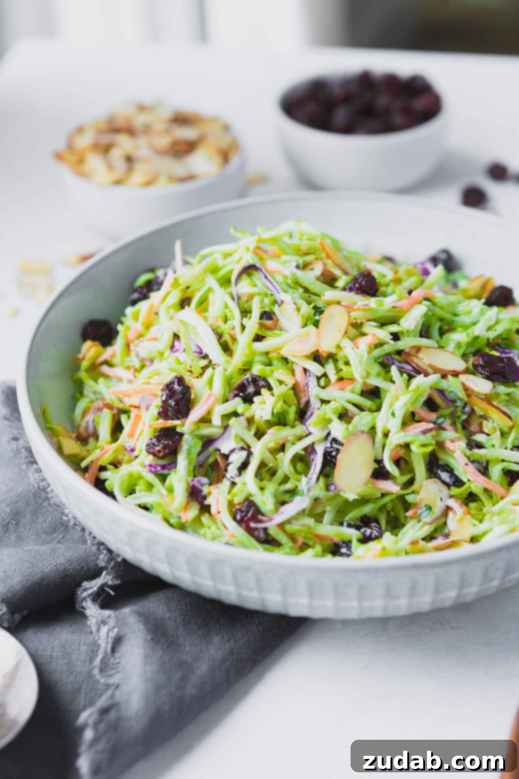 A photo of a big white ceramic bowl full of broccoli slaw topped with raisins and sliced almonds.