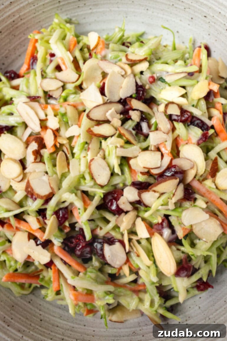 A large ceramic bowl filled with creamy broccoli slaw, featuring shredded broccoli stems, carrots, craisins, and sliced almonds, coated in a homemade dressing.