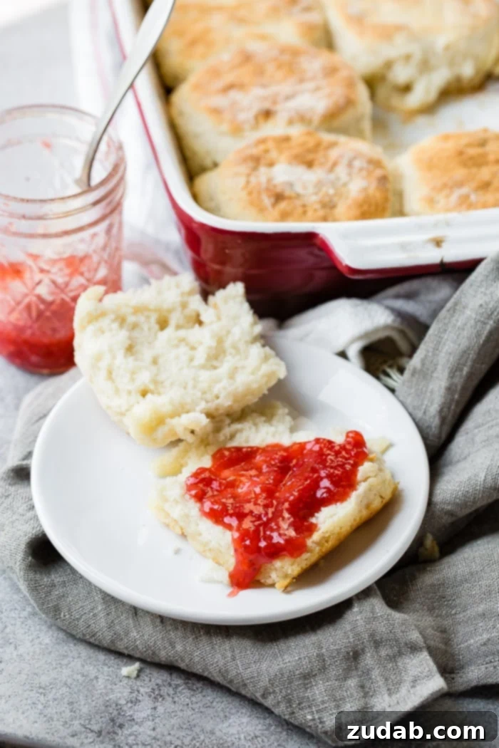 Surprisingly Easy 7 Up Biscuits 5 a gray napkin with a white plate, cut open 7 up biscuit with strawberry jam and a jar of ham and pan of biscuits behind it