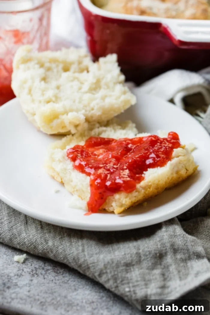 Surprisingly Easy 7 Up Biscuits 9 a gray napkin with a white plate, cut open 7 up biscuit with strawberry jam and a jar of ham and pan of biscuits behind it