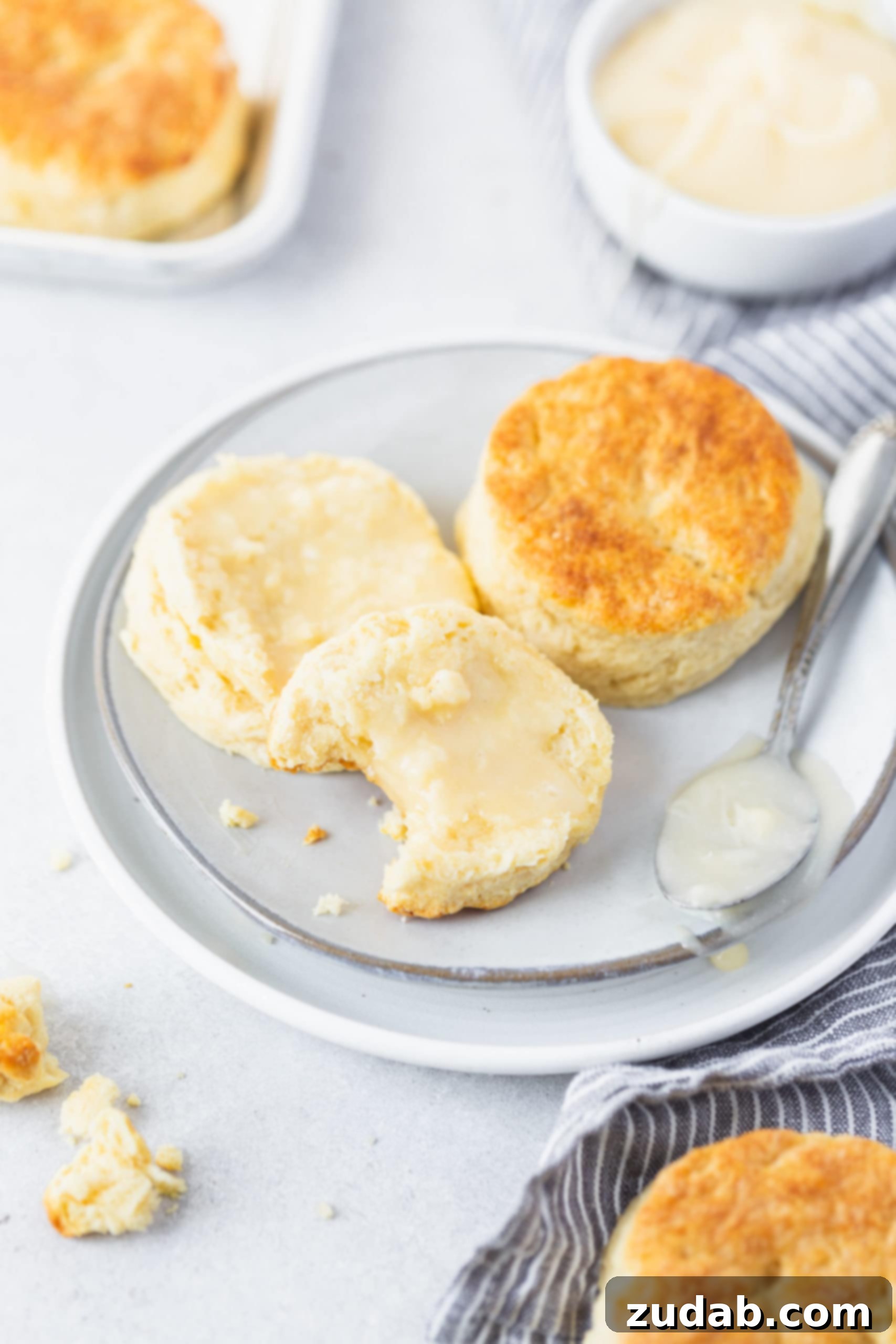 Buttermilk Biscuits Two buttermilk biscuits on a white plate. One biscuit is cut in half and spread with honey butter. A bite has been taken out of one biscuit half.