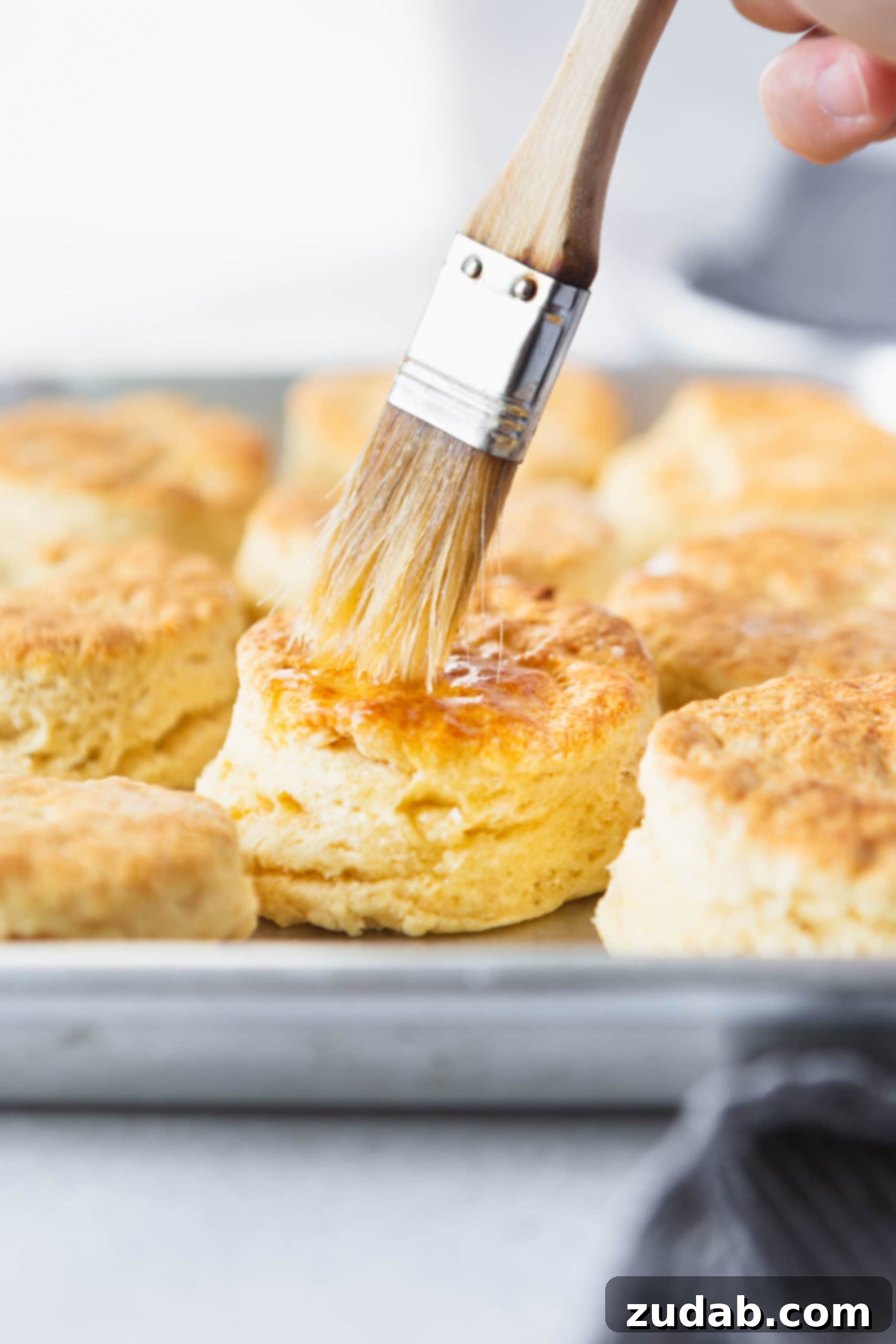 Buttermilk Biscuits A pan full of golden brown buttermilk biscuits. Butter is being brushed on the top of a biscuit with a pastry brush.
