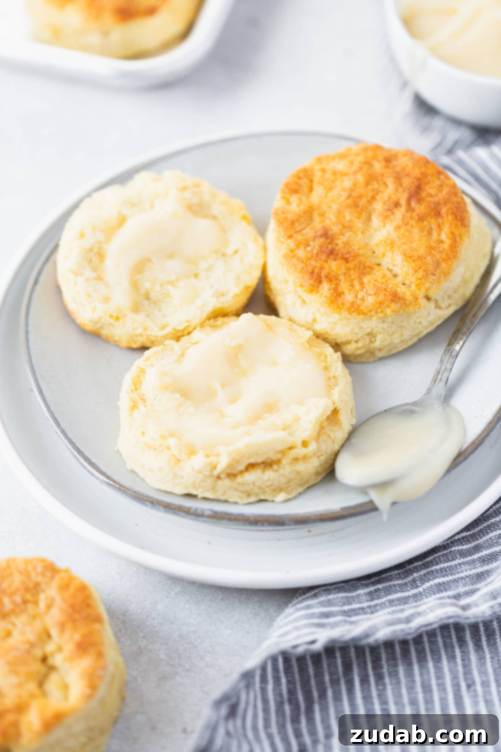 Buttermilk Biscuits Two buttermilk biscuits on a small white plate. One biscuit is broken in half and has been buttered.