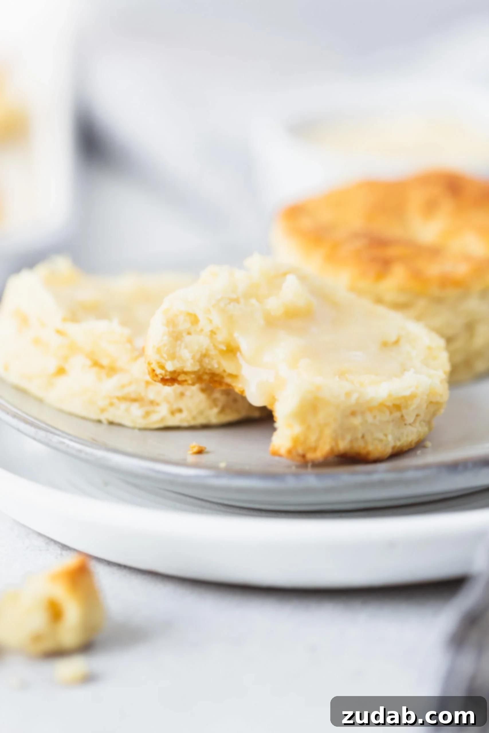 Buttermilk Biscuits Two buttermilk biscuits on a plate. One biscuit is broken in half and has been spread with honey butter. A bite has been taken from one of the halves.