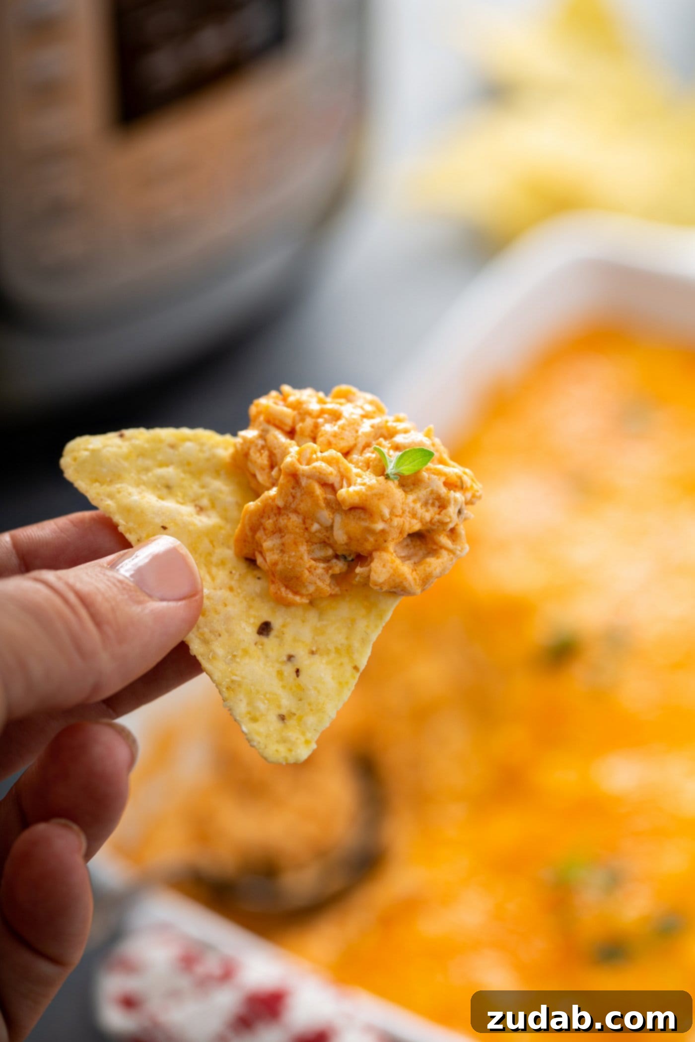 Instant Pot Buffalo Chicken Dip A tortilla chip that has been dipped in buffalo chicken dip. The casserole dish of dip is in the background and you can see part of an instant pot.