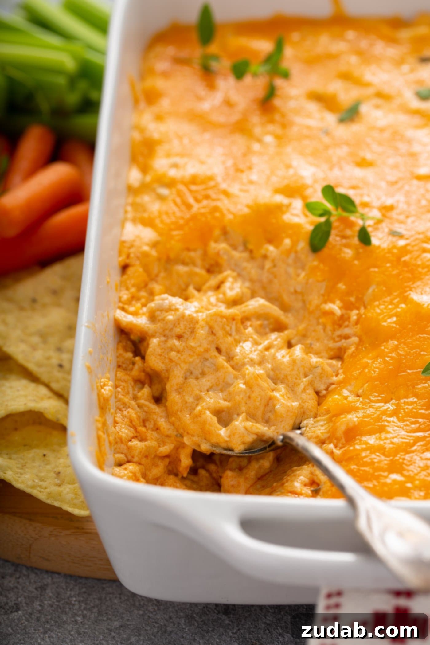 Instant Pot Buffalo Chicken Dip A casserole dish of buffalo chicken dip. A spoon is lifting a scoop of dip. There is shredded chicken in buffalo sauce with melted cheese on top. A few fresh parsley leaves are sprinkled on top. Chips and carrots are next to the dish of dip.