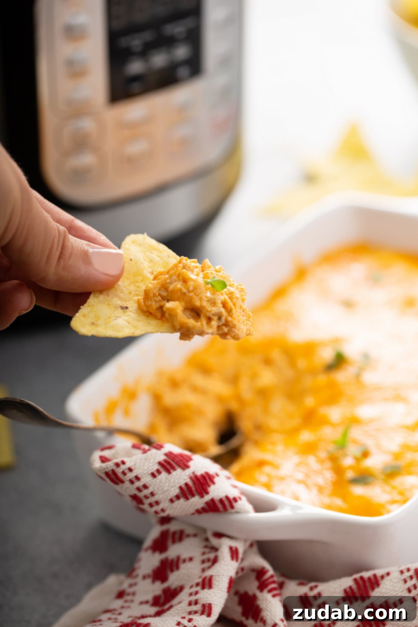 Instant Pot Buffalo Chicken Dip A chip dipped in buffalo chicken dip. There is shredded chicken and melted cheese on the chip and a dish of buffalo chicken dip is beneath the chip. More chips are beside the dish.