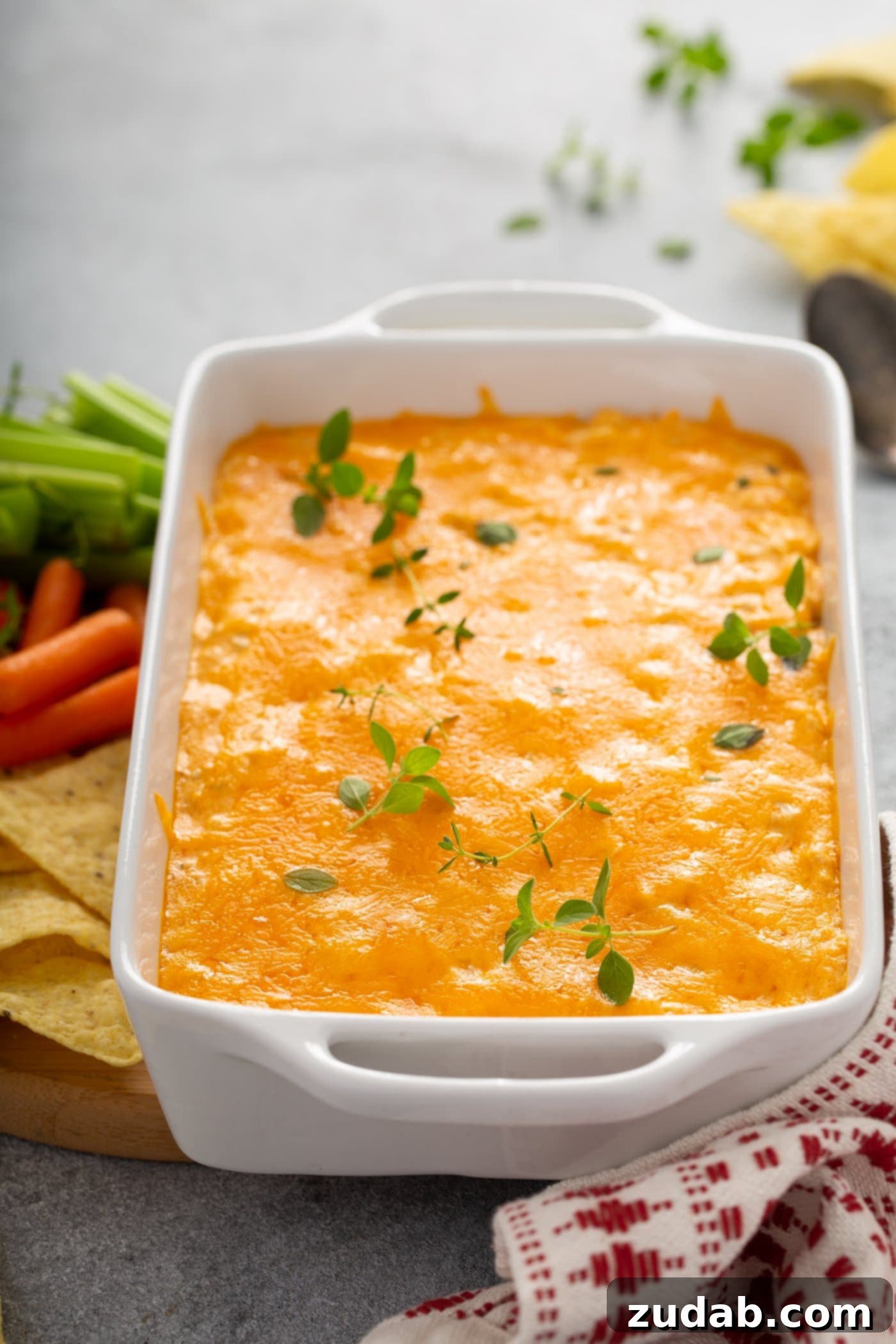 Instant Pot Buffalo Chicken Dip A casserole dish filled with buffalo chicken dip. It is covered with melted cheese and fresh parsley leaves are sprinkled on top. There are chips, carrots, and celery next to the dish of dip.