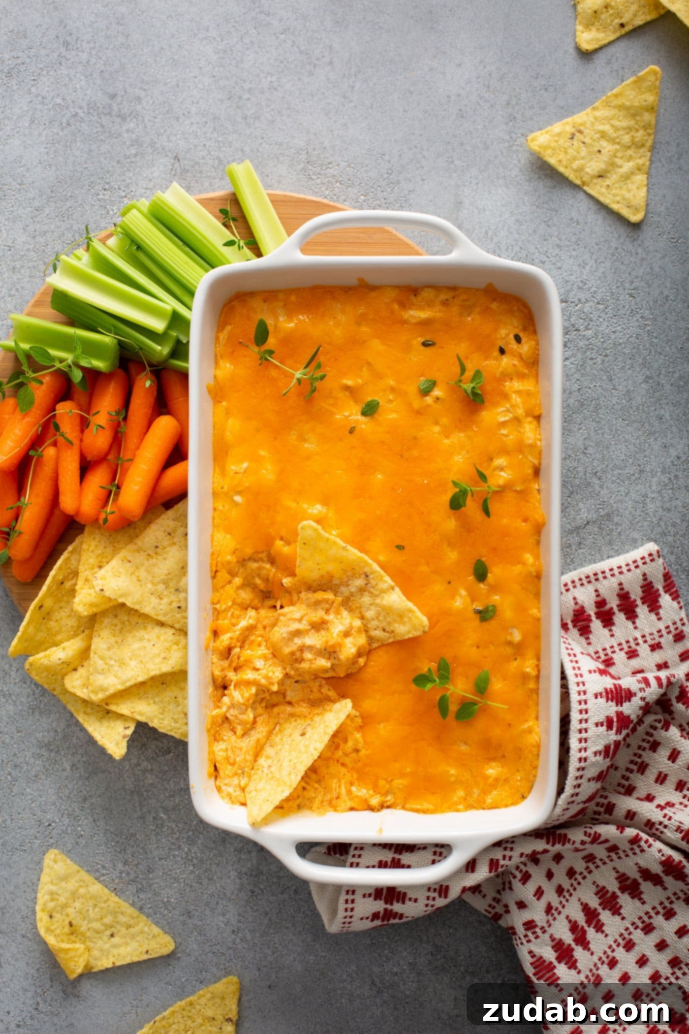 Instant Pot Buffalo Chicken Dip A casserole dish of instant pot buffalo chicken dip. There are two chips that have been dipped into the cheesy dip. Chips, carrots, and celery are next to the dip and a red and white checked tea towel is next the the dish.