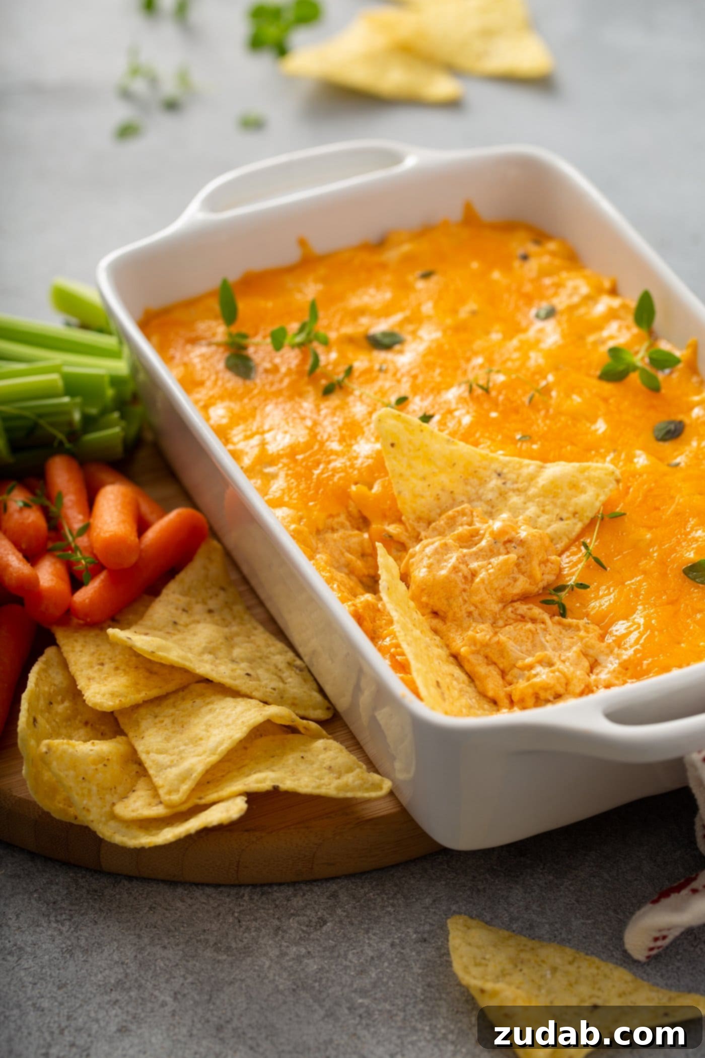Instant Pot Buffalo Chicken Dip A casserole dish with buffalo chicken dip. A scoop has be taken out of the dip and there are a couple of chips laying in the dip. The dip is covered in melted cheese and sprinkled with parsley leaves. There are chips, carrots and celery laying next to the dip.