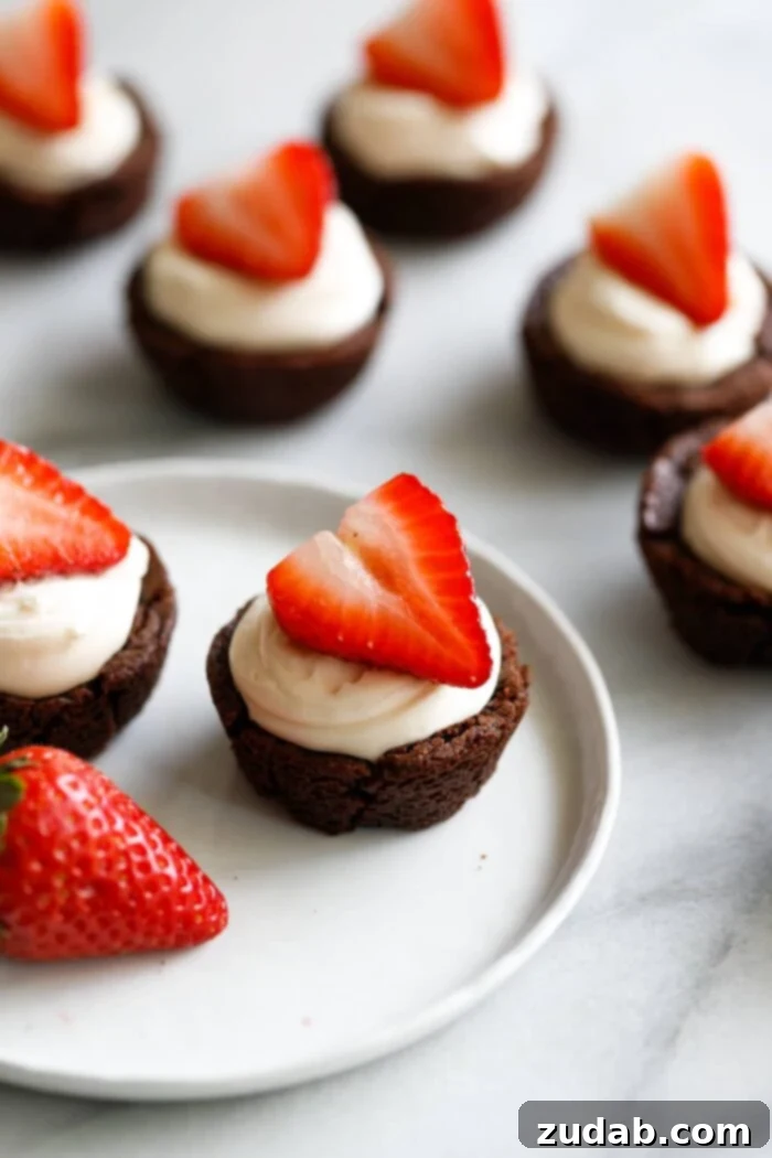 Black Tie Brownie Treats 5 A marble background with a white plate filled with tiny brownie cups filled with a white chocolate cream cheese filling and a sliced strawberry on top making these the cutest tuxedo brownie cups