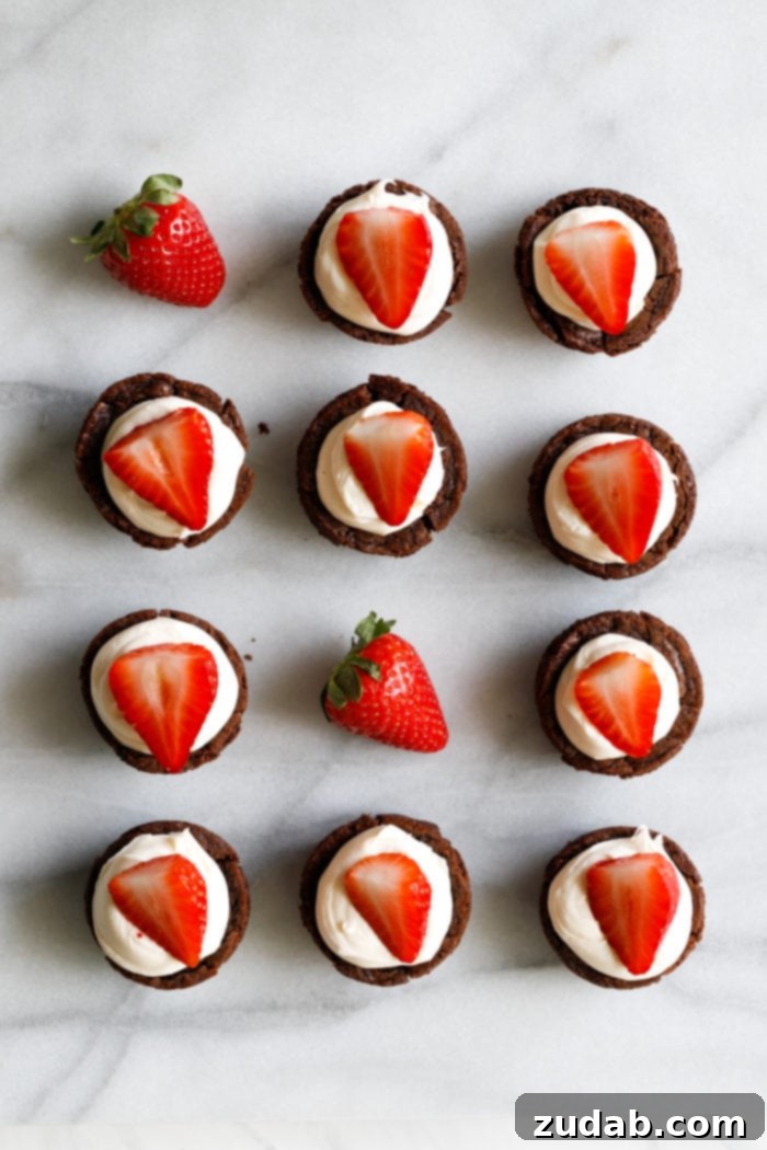 Black Tie Brownie Treats 6 A marble background with a white plate filled with tiny brownie cups filled with a white chocolate cream cheese filling and a sliced strawberry on top making these the cutest tuxedo brownie cups