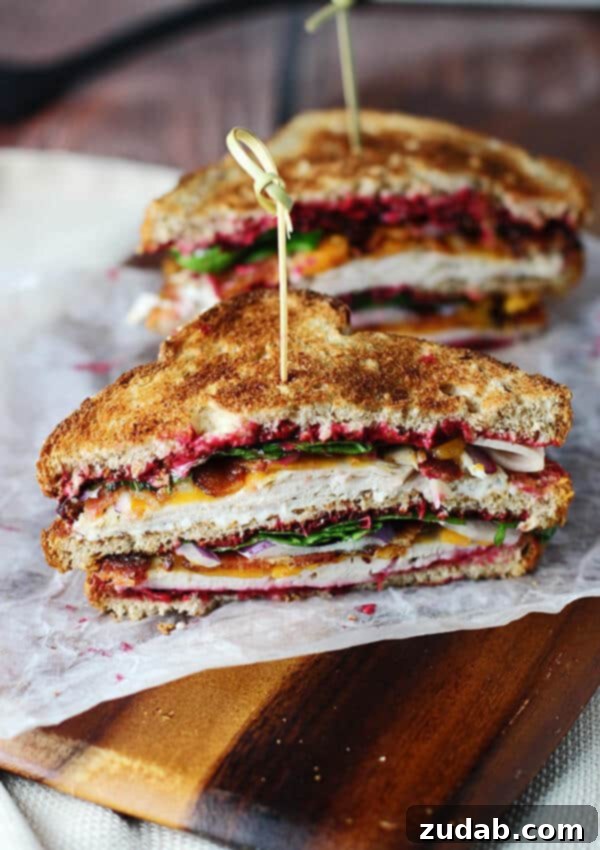 Award-Winning Instant Pot Turkey: The Ultimate Recipe 8 A mouth-watering photo of a gourmet sandwich, sliced in half, showcasing its rich layers. Toasted bread encases tender turkey, sweet cranberry sauce, melted cheddar cheese, crisp lettuce, and savory bacon. One half of the sandwich is prominently displayed in the foreground, revealing its delectable profile, while the other rests softly in the background. The sandwich is presented on a piece of parchment paper atop a rustic wooden table, inviting viewers to indulge.