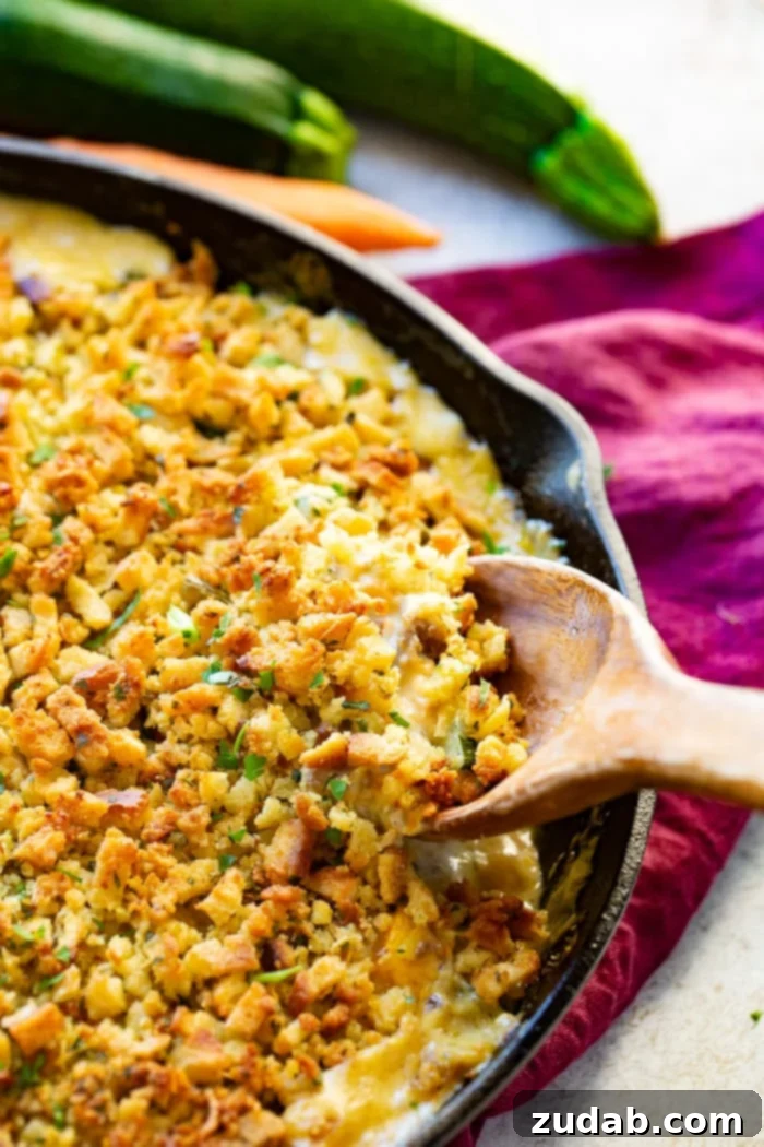 Hearty Creamy Zucchini Sausage Bake 2 A cast iron skillet overflowing with creamy zucchini sausage casserole, topped with golden-brown stuffing, being served with a wooden spoon. The skillet rests on a purple cloth napkin, surrounded by fresh zucchinis and a carrot, hinting at its garden-fresh origins.