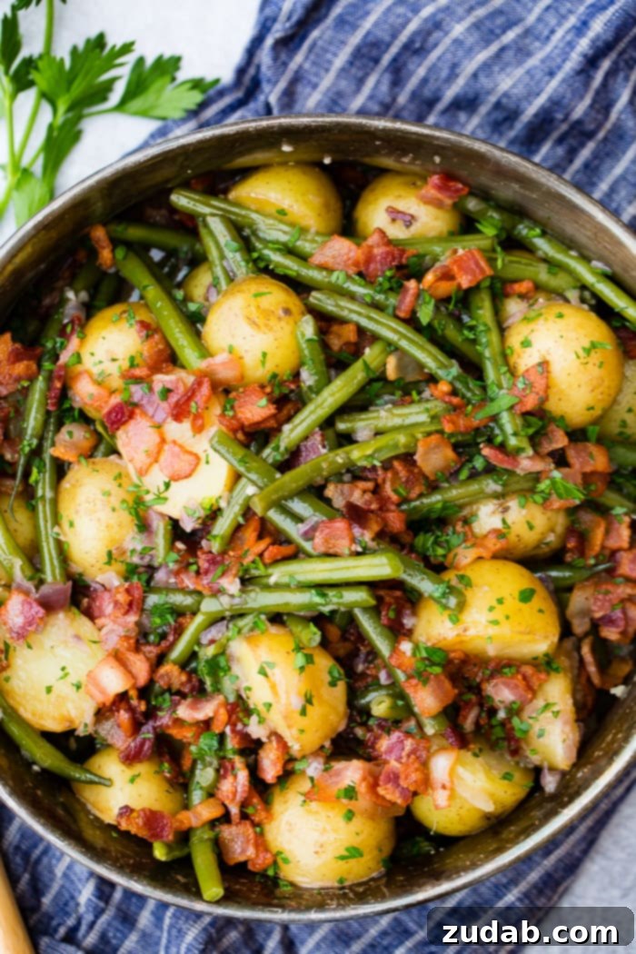Effortless Instant Pot Green Beans and Potatoes 2 A silver serving dish sitting on a blue cloth napkin that is full of cooked fresh green beans, potatoes, and bacon pieces sprinkled with fresh parsley.