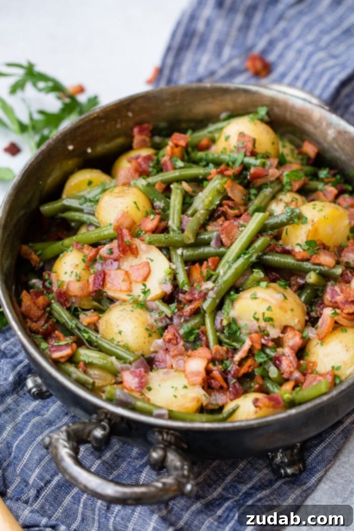 Effortless Instant Pot Green Beans and Potatoes 3 A photo of a silver serving dish sitting on a blue cloth napkin that is full of cooked fresh green beans, potatoes, and bacon pieces sprinkled with fresh parsley.