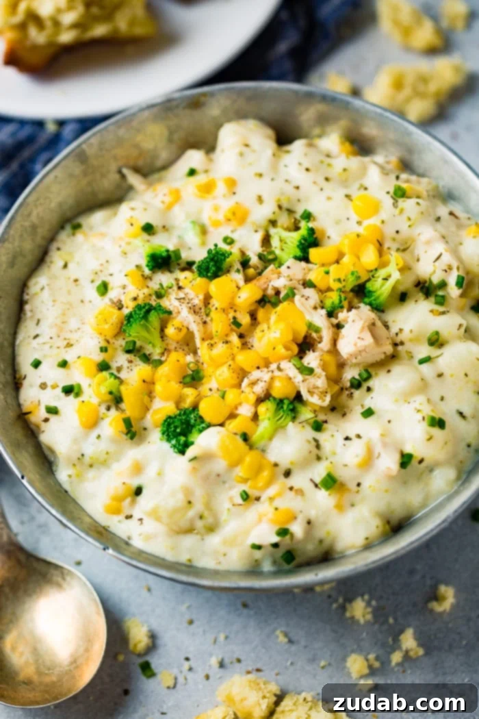 Hearty Chicken & Garden Chowder 4 A close-up of a silver bowl filled with rich Chicken Potato Broccoli Corn Chowder, garnished with fresh chives, set on a grey wooden surface beside a blue striped towel.