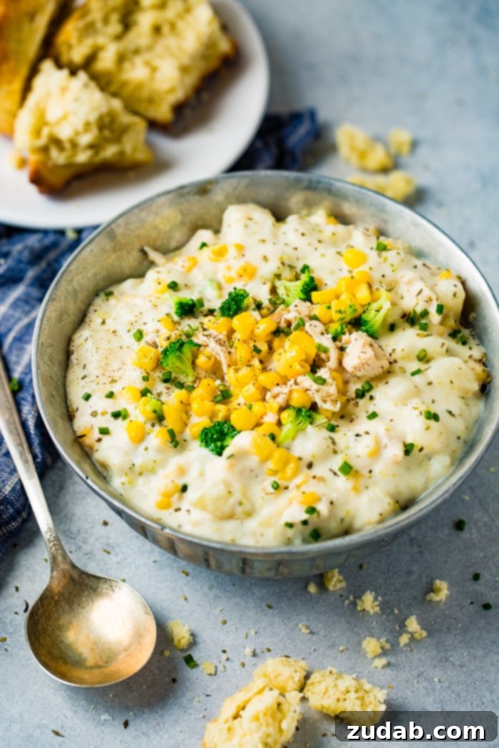Hearty Chicken & Garden Chowder 5 A silver bowl of creamy Chicken Potato Broccoli Corn Chowder, garnished with fresh chives, on a grey backdrop with a blue towel, highlighting the inviting warmth of the soup.