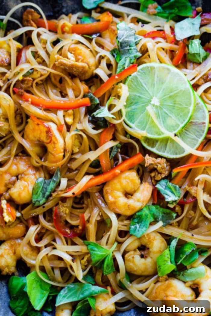 Zesty One-Pan Asian Shrimp Noodles 3 A close-up view of succulent cooked shrimp nestled among tender rice noodles, vibrant red bell peppers, and fragrant fresh basil leaves, all on a blue plate with lime wedges.