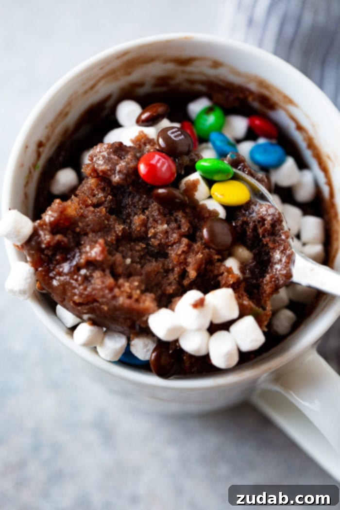 A white mug filled with a warm, baked brownie, generously adorned with mini marshmallows and colorful mini M&Ms. A spoon is taking a delectable scoop from the brownie, highlighting its soft, gooey texture and the delightful mix of toppings.