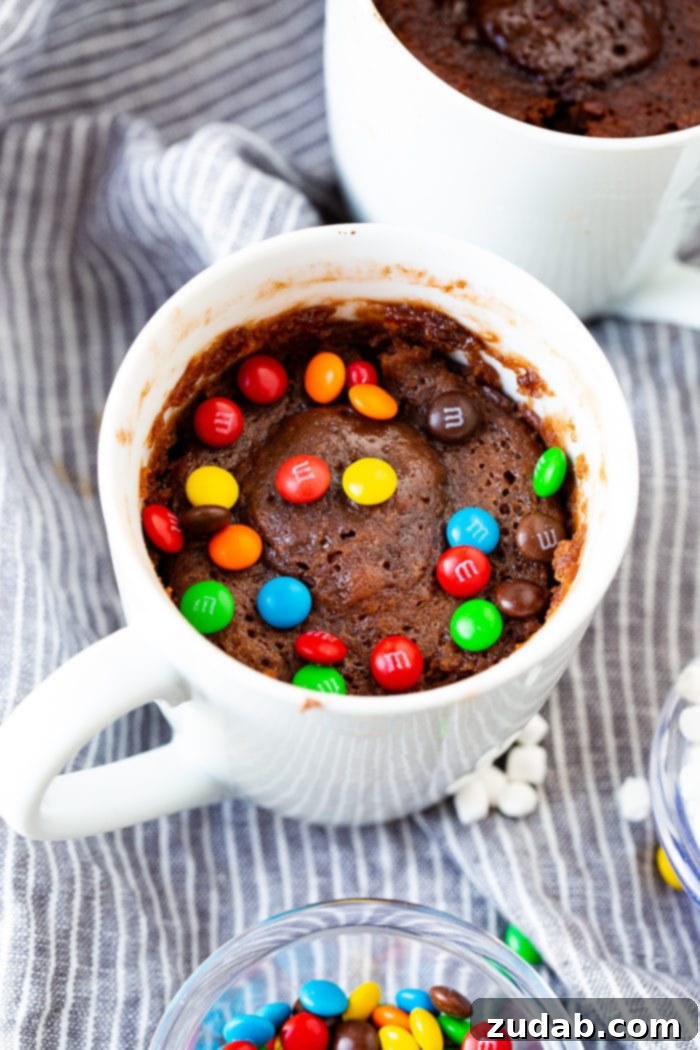 A close-up shot of a white mug brimming with a warm, fudgy brownie, beautifully decorated with a generous scattering of colorful mini M&Ms. The vibrant candies add a playful touch to this comforting, single-serving dessert.