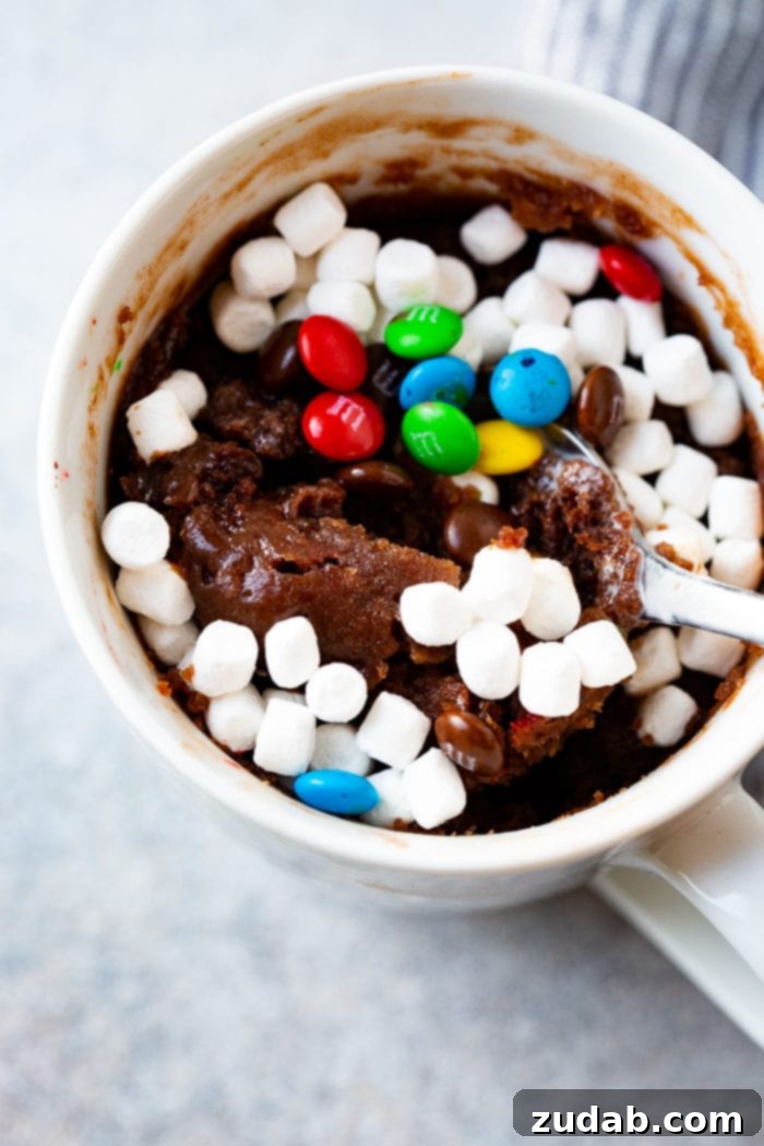 A close-up of a white mug containing a baked brownie, topped with a delightful mix of mini marshmallows and colorful mini M&Ms. A spoon is mid-scoop, capturing the moment of indulgence from this warm, single-serving chocolate dessert.