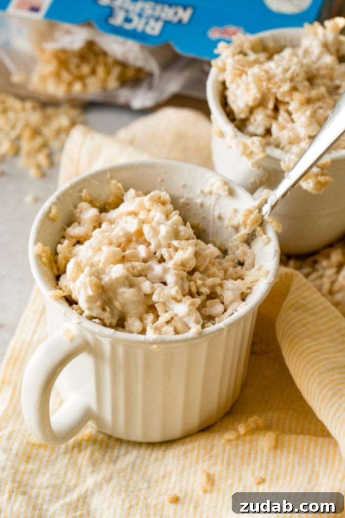 2-Minute Rice Krispie Treat in a Mug 4 A photo of a white mug full of rice krispies treat with a spoon in it on an orange cloth napkin and another mug of rice krispies in the background.