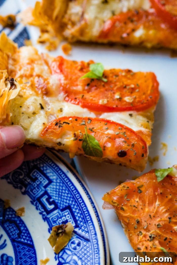 Country Tomato Galette 5 A hand carefully holds a slice of the rustic tomato tart, rich with baked tomatoes, cheeses, and basil, poised over a blue and white plate, ready to be enjoyed.