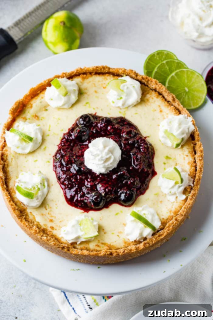 Sunshine Lime Cheesecake with Berry Swirl 2 An aerial photo of a whole cheesecake on a white serving platter with dollops of whipped cream around the edges with a slice of lime in each dollop and a pile of triple berry sauce in the middle with a dollop of whipped cream on top