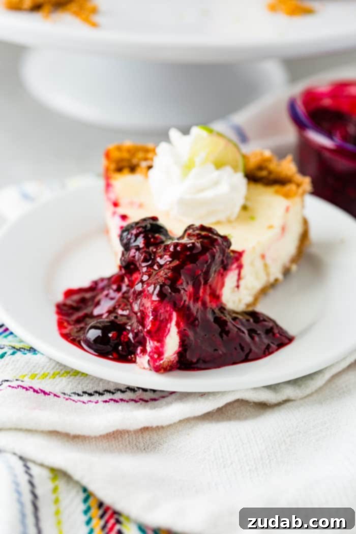 Sunshine Lime Cheesecake with Berry Swirl 11 A photo of a slice of lime cheesecake on a while plate covered in triple berry sauce and dollop of whipped cream and a slice of lime.