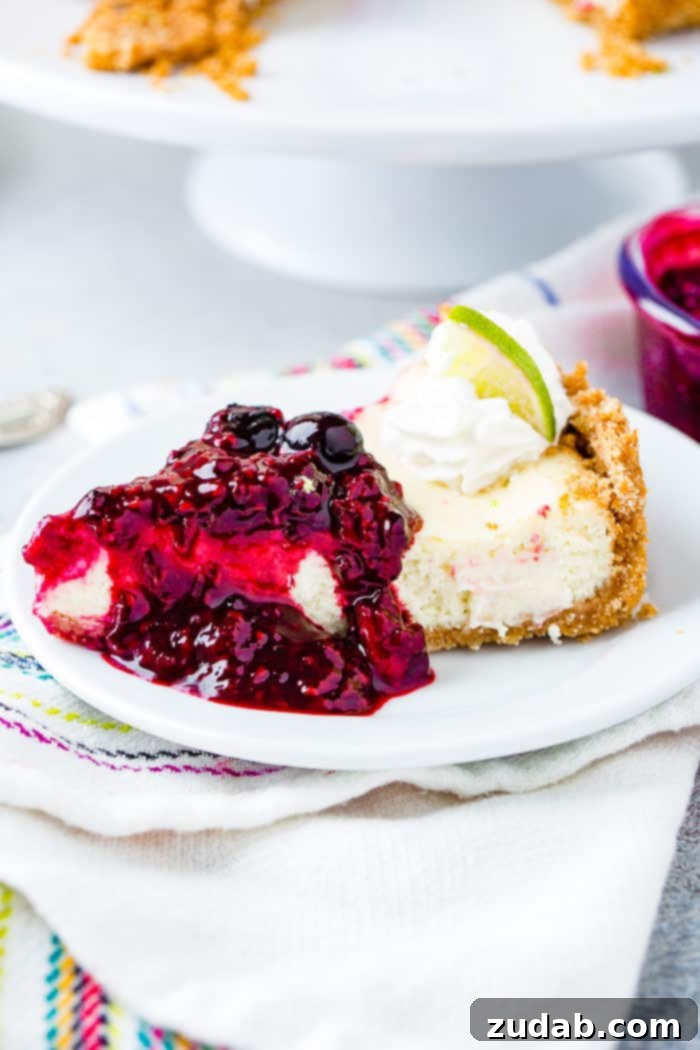 Sunshine Lime Cheesecake with Berry Swirl 5 A photo of a slice of lime cheesecake on a while plate covered in triple berry sauce and dollop of whipped cream and a slice of lime.