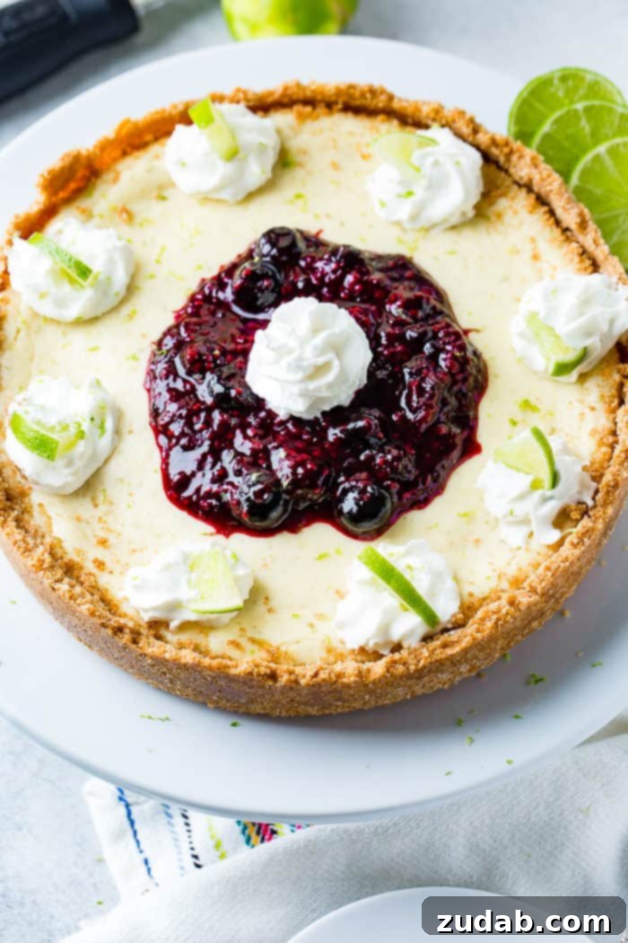 Sunshine Lime Cheesecake with Berry Swirl 7 An aerial photo of a whole cheesecake on a white serving platter with dollops of whipped cream around the edges with a slice of lime in each dollop and a pile of triple berry sauce in the middle with a dollop of whipped cream on top