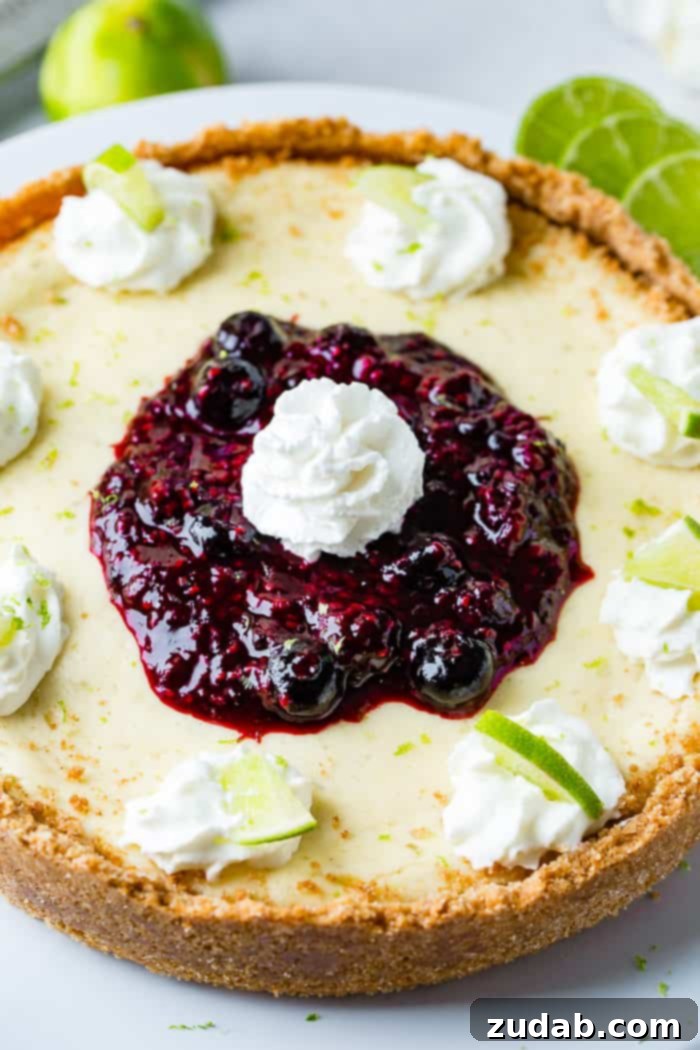 Sunshine Lime Cheesecake with Berry Swirl 9 A zoomed in aerial photo of a whole cheesecake on a white serving platter with dollops of whipped cream around the edges with a slice of lime in each dollop and a pile of triple berry sauce in the middle with a dollop of whipped cream on top