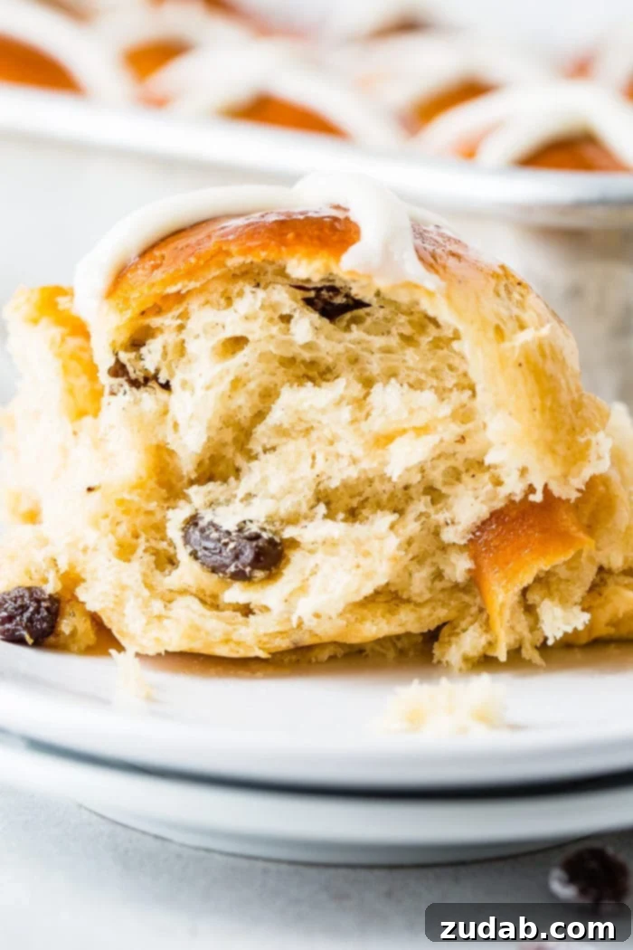 Pillowy Soft Hot Cross Buns 4 A photo of a cross section of a hot cross bun sitting on a white plate full of raisins and topped with a white frosting cross.