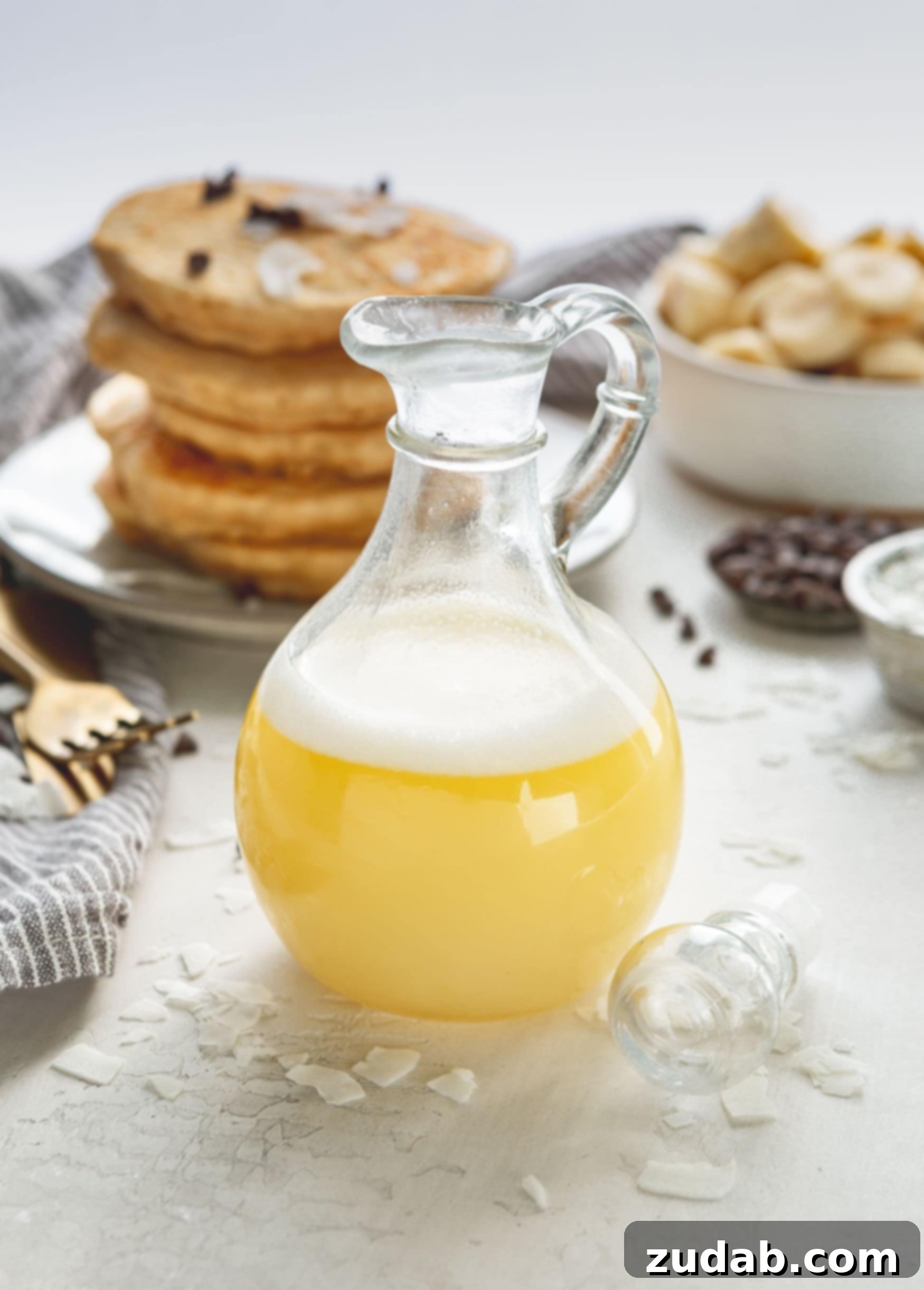 Luscious Coconut Buttermilk Syrup 2 A syrup container filled with light yellow coconut buttermilk syrup, topped with a delicate white foam. In the blurred background, a stack of golden pancakes awaits, adorned with scattered chocolate chips.