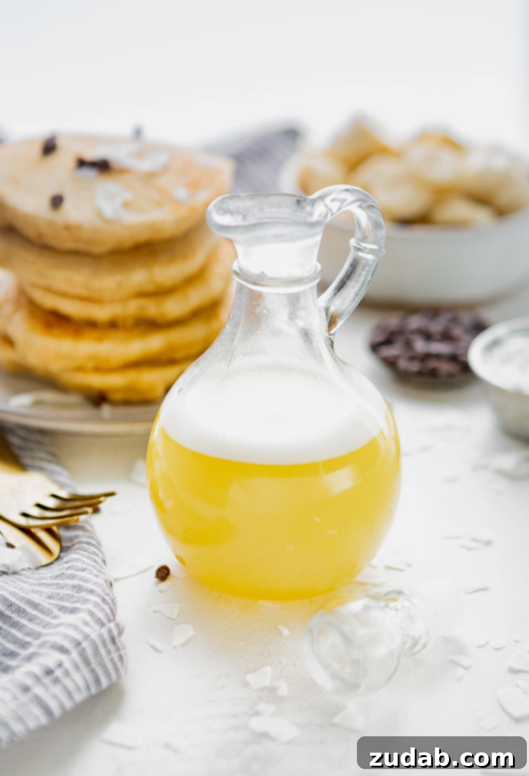 Luscious Coconut Buttermilk Syrup 3 A small syrup pitcher gracefully pours light yellow coconut buttermilk syrup, adorned with a delicate white foam. In the background, a tempting stack of chocolate chip pancakes and a bowl of fresh sliced bananas complete a delicious breakfast scene.