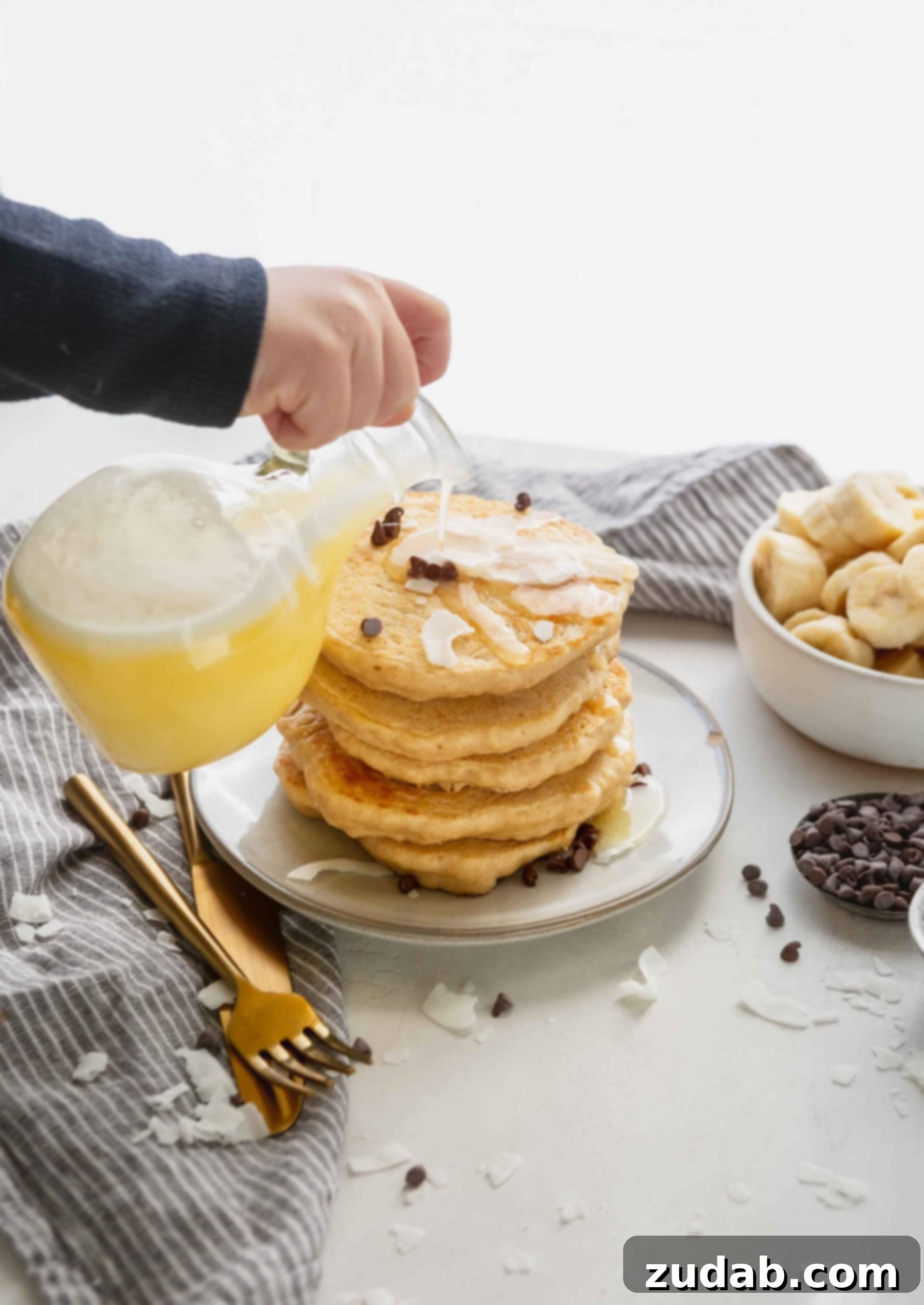 Luscious Coconut Buttermilk Syrup 4 A stack of golden brown pancakes being generously drenched with light yellow coconut buttermilk syrup, showing a frothy top. A bowl of sliced bananas and scattered chocolate chips are arranged beautifully around the pancakes.