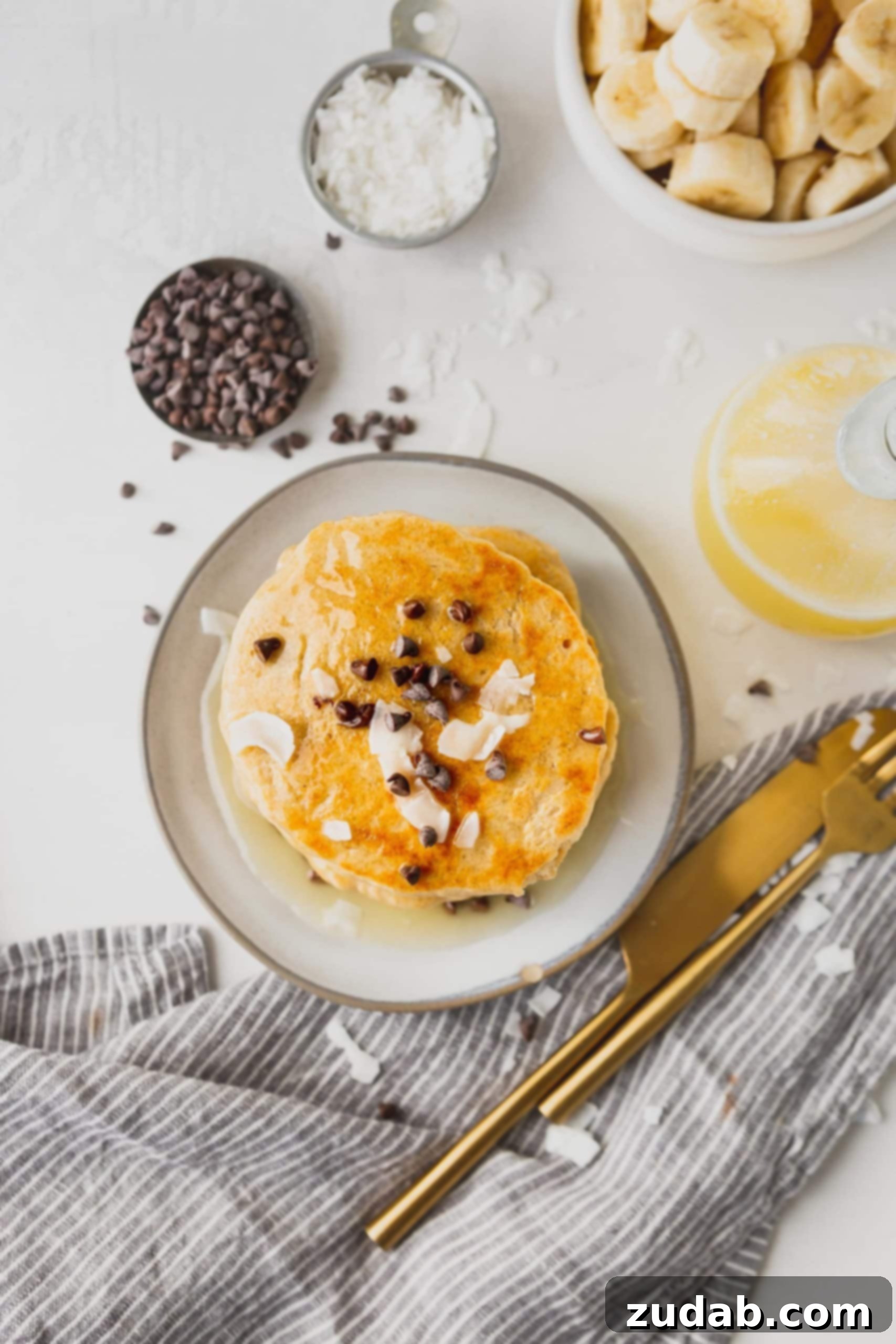 Luscious Coconut Buttermilk Syrup 5 A top-down view of a delectable stack of pancakes topped with chocolate chips and delicate coconut flakes. A small container of chocolate chips and a syrup pitcher filled with coconut buttermilk syrup are neatly placed beside the pancakes.