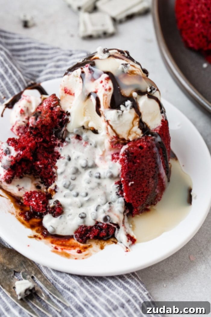 A close-up shot of a single red velvet molten lava cake, revealing its rich, dark red crumb. The top is generously covered with a glistening scoop of vanilla ice cream, and a decadent chocolate sauce cascades down its sides, pooling at the base.