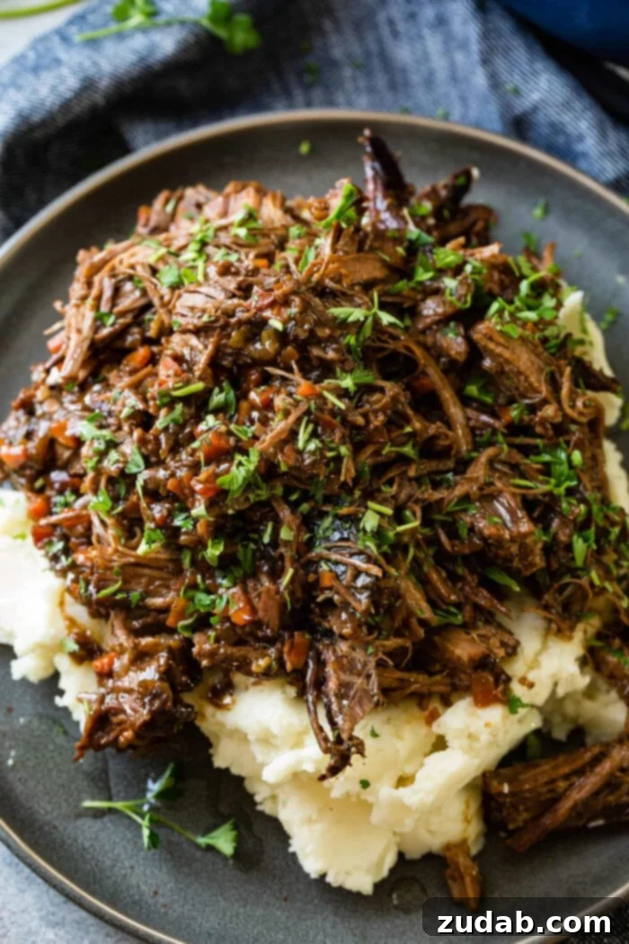 Hearty Oven-Braised Pot Roast 2 A photo of a dark gray plate with a pile of mashed potatoes on it covered in juicy shredded braised beef topped with chopped fresh parsley.