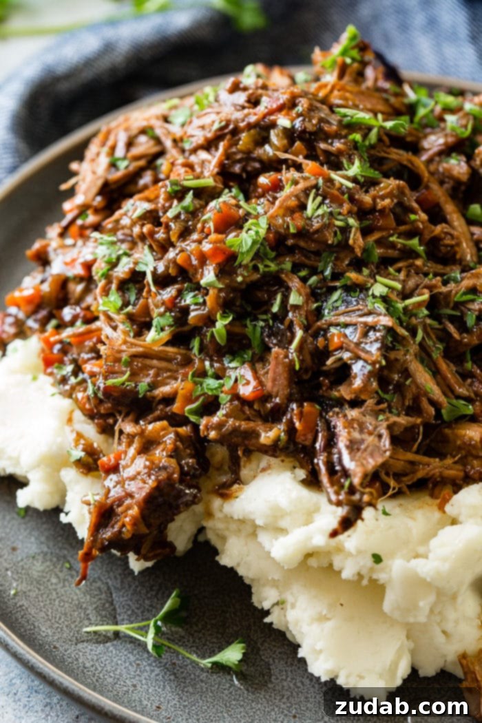 Hearty Oven-Braised Pot Roast 3 A close up photo of a dark gray plate with a pile of mashed potatoes on it covered in juicy shredded braised beef topped with chopped fresh parsley.