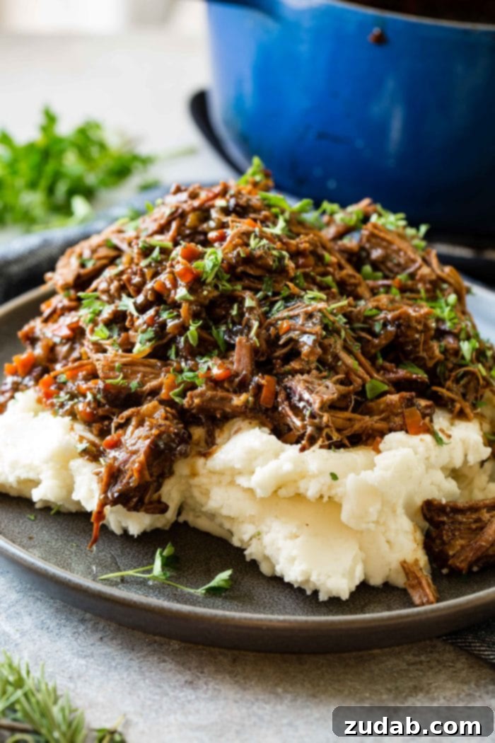 Hearty Oven-Braised Pot Roast 4 A photo of a dark gray plate with a pile of mashed potatoes on it covered in juicy shredded braised beef topped with chopped fresh parsley.
