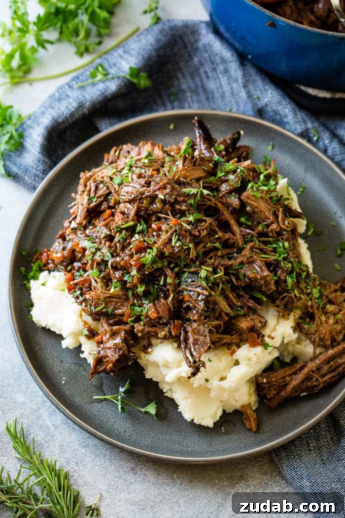 Hearty Oven-Braised Pot Roast 8 A photo of a dark gray plate with a pile of mashed potatoes on it covered in juicy shredded braised beef topped with chopped fresh parsley.