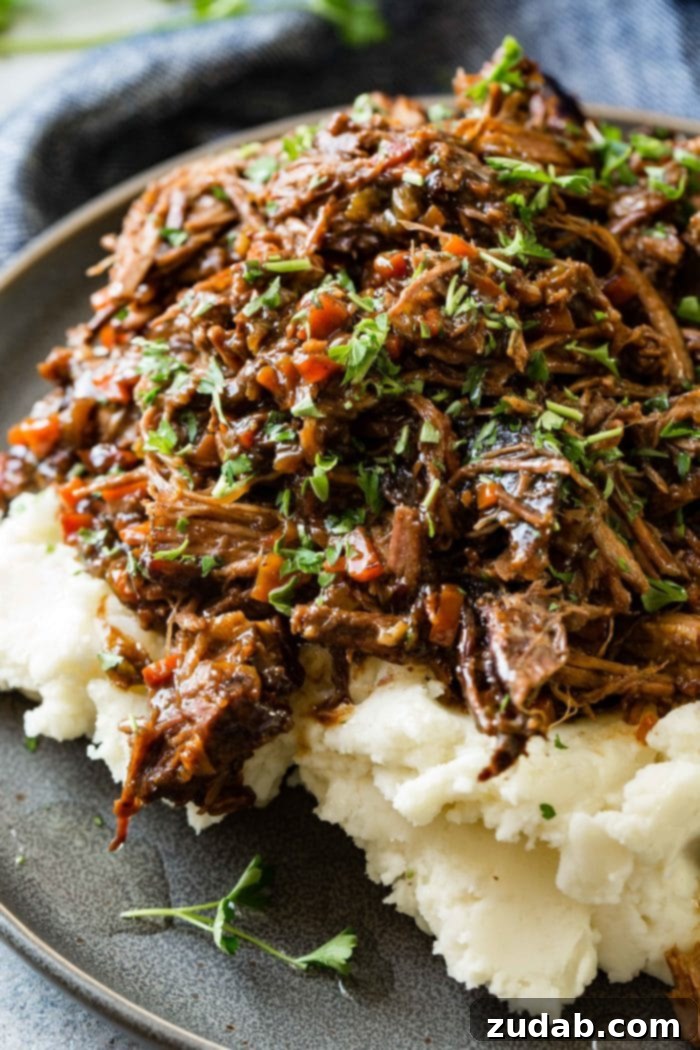 Hearty Oven-Braised Pot Roast 9 A photo of a dark gray plate with a pile of mashed potatoes on it covered in juicy shredded braised beef topped with chopped fresh parsley.