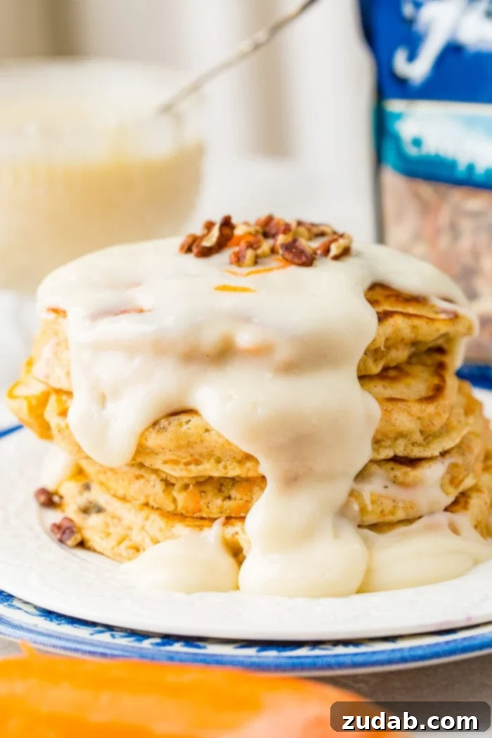 Sweet and Spiced Carrot Cake Pancakes 2 A photo of a stack of four carrot cake pancakes with a thick white cream cheese sauce poured over the top with a few chopped pecans and shredded carrots on top.