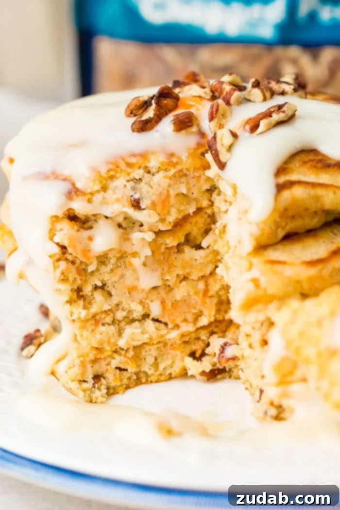 Sweet and Spiced Carrot Cake Pancakes 5 A photo of a stack of four carrot cake pancakes with a slice taken out with a thick white cream cheese sauce poured over the top with a few chopped pecans and shredded carrots on top.