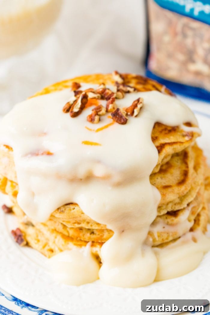 Sweet and Spiced Carrot Cake Pancakes 6 A photo of a stack of four carrot cake pancakes with a thick white cream cheese sauce poured over the top with a few chopped pecans and shredded carrots on top.